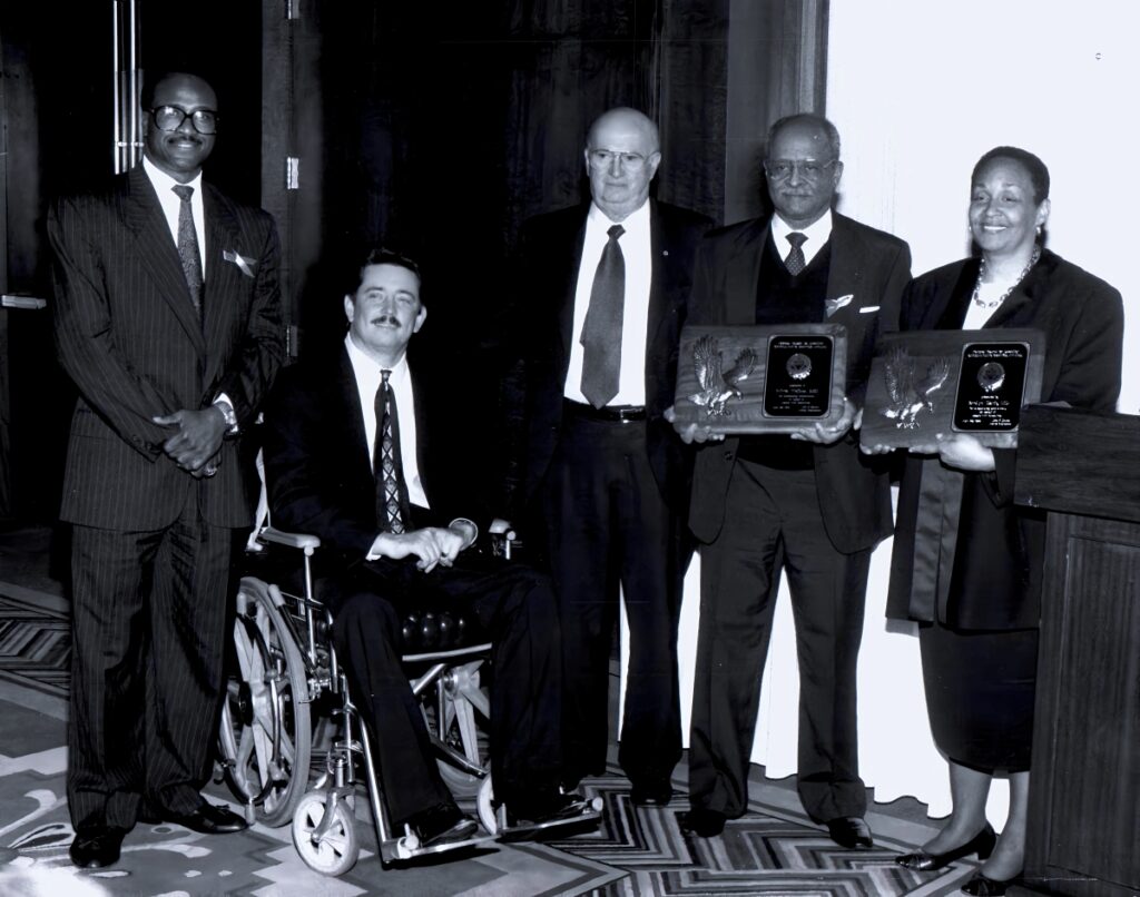 A black and white picture of 5 people, holding an award with one person in a wheelchair