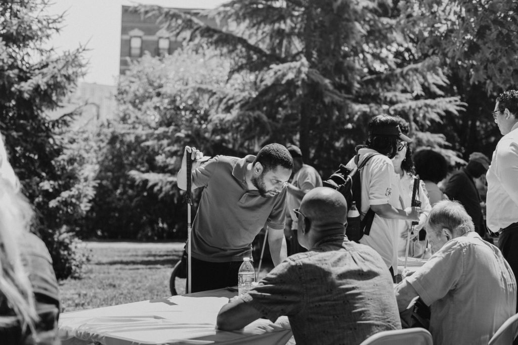 A black and white picture of a consumer with a white cane talking with two visually impaired gentlemen. The two gentlemen are sitting at a table at an event in a park.